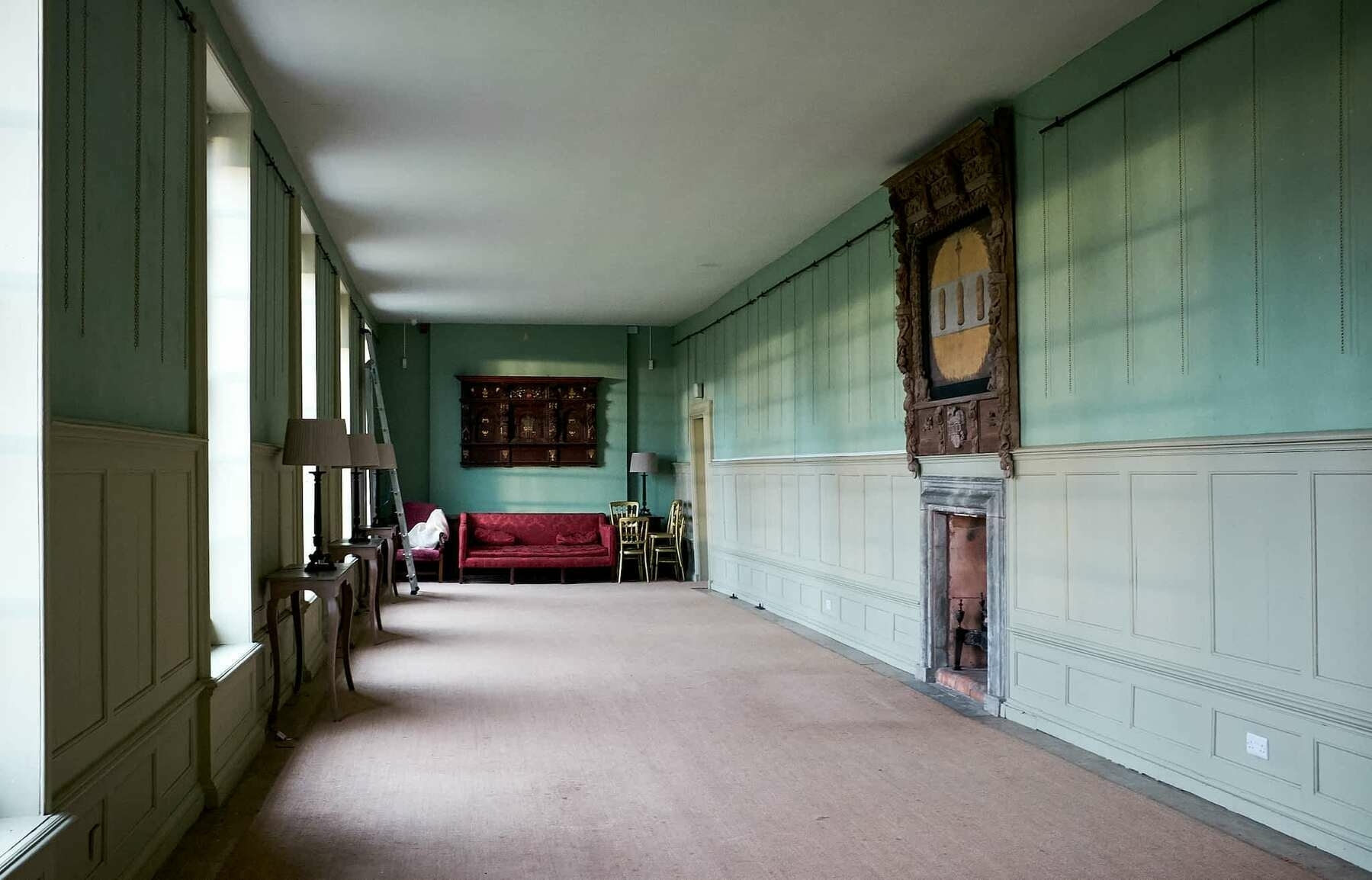 A view down a long empty gallery room. Winter light enters from windows on the left. The walls are painted a soft robin's egg blue colour, and there is a red sofa at the far end of the room.